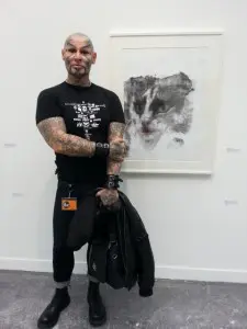 L'artiste français Jean-Luc Verna, posant, en pied et en peau tatouée, devant l'une de ses oeuvres à la galerie Air de Paris: "Pale Blue Eyes" (2015), dessin à l'encre et au produit de maquillage. L'étrangeté et la félinité... 